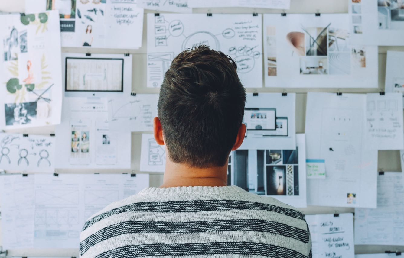 A man stands in front of a wall covered in various papers, deep in thought or contemplation.