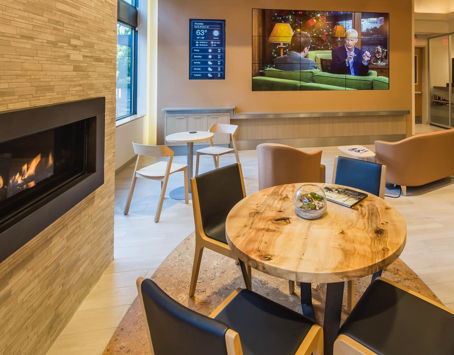 A waiting room with a wood table, black chairs, a wall of televisions and a fireplace.