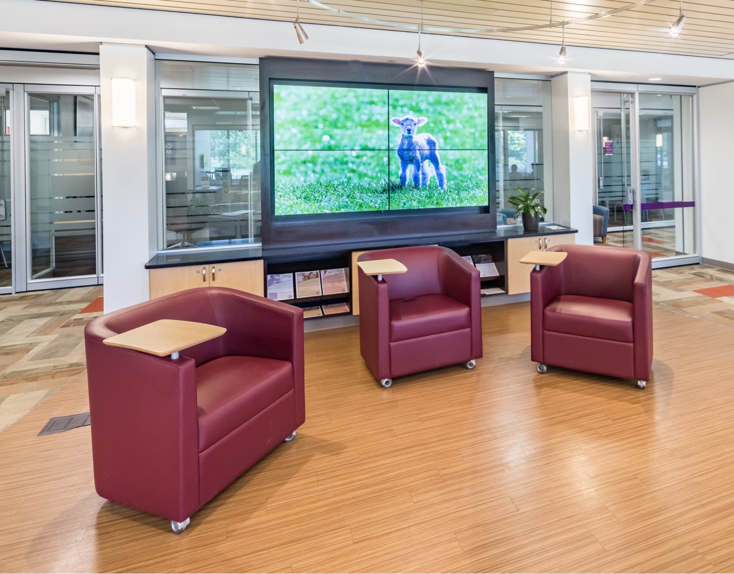 Three red, leather chairs with attached tables, sitting in front of wall of television screens.