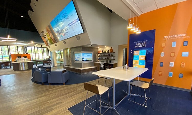 A bank lobby with a waiting room with upholstered chairs, and a high table with wood chairs.