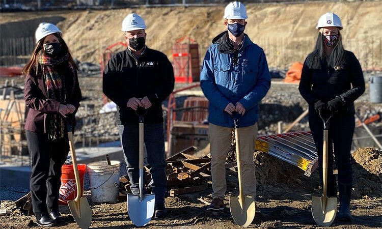 Four people wearing masks and hardhats, standing in a row, each holding a shovel.