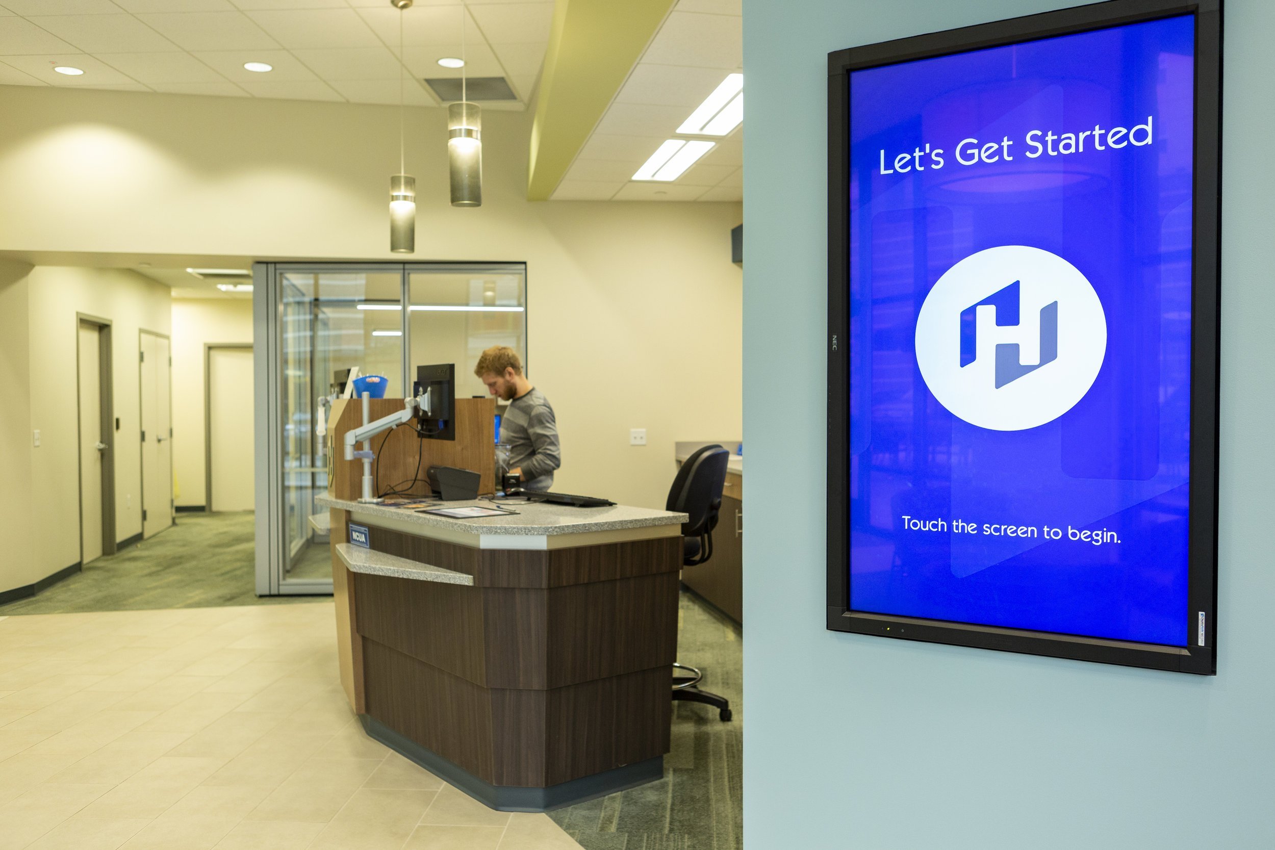 A screen on a wall, with the words "let's get started", with a man working at a desk in the background.