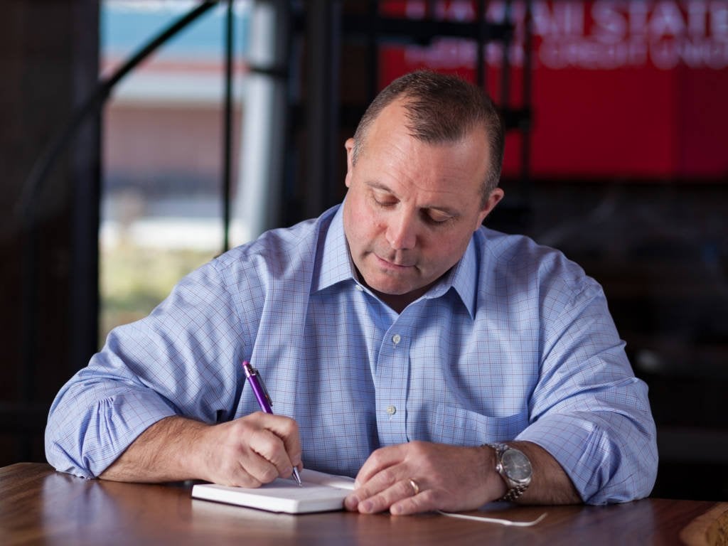 A man in a blue button-up writing on a pad of paper with a purple pen.