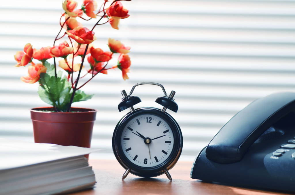 A table with an alarm clock, landline phone, and a flower pot in the background.