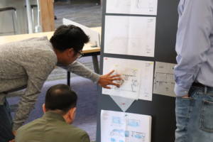 Three men looking at blueprints, hung on a black wall.