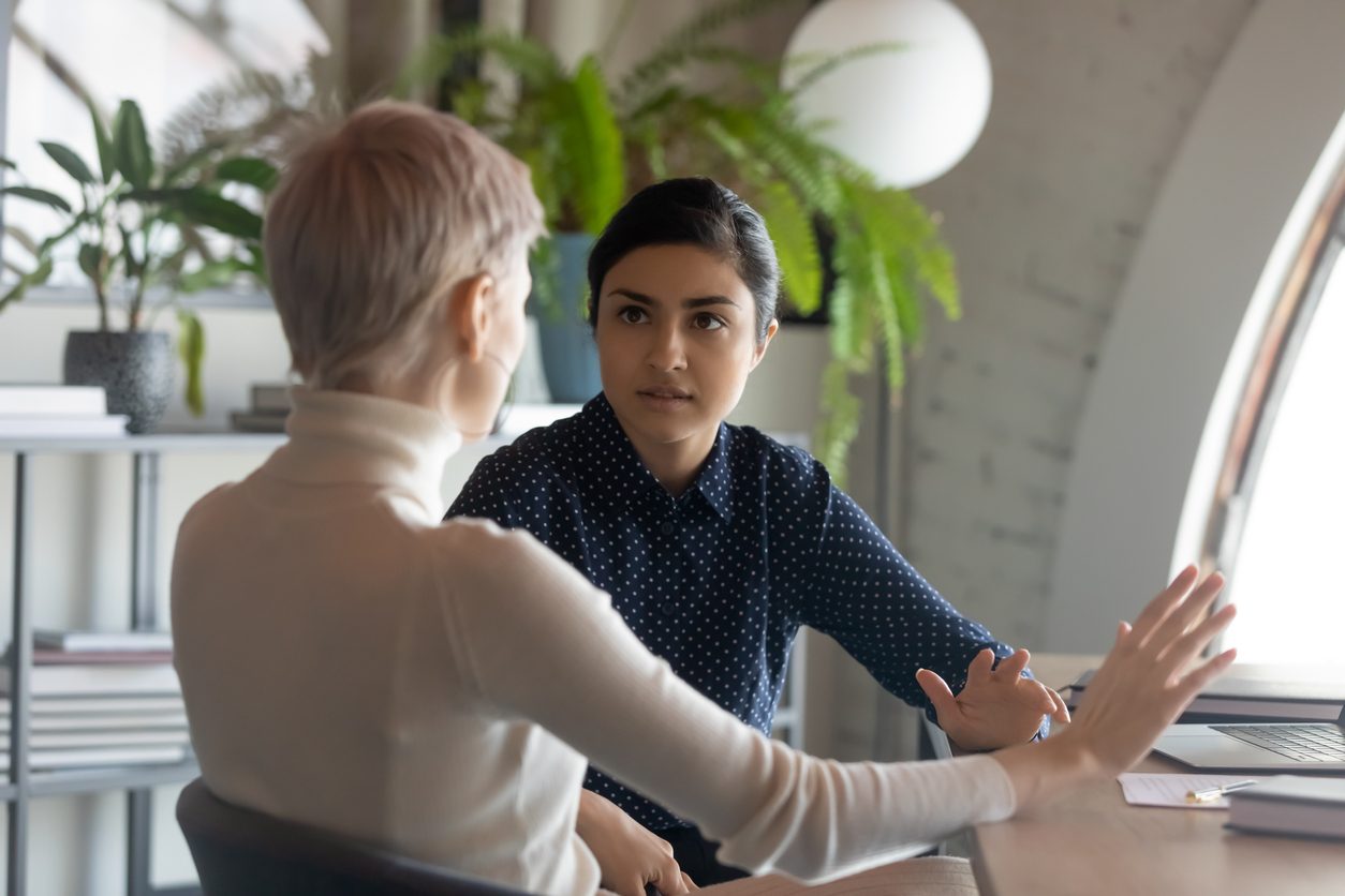 Two women engaged in conversation in a professional office environment, discussing work-related matters.