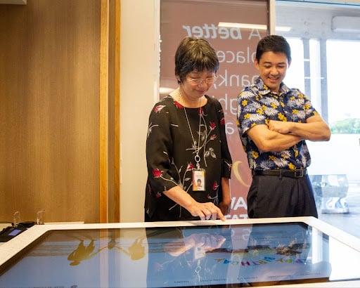 A woman and man engage with a large digital touchscreen display in a modern credit union lobby for an interactive banking customer experience.