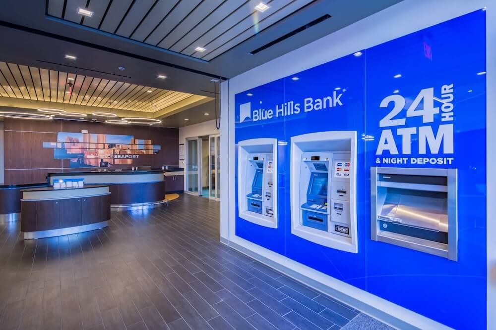 The image showcases the interior of a modern bank branch featuring a distinct Blue Hills Bank branding. The design includes sleek, contemporary furniture and a reception desk with a mural of a cityscape in the background. Prominently displayed are two ATM machines housed in a vibrant blue wall with clear signage indicating “24 Hour ATM & Night Deposit” facilities. This setting highlights the bank’s commitment to customer convenience and accessibility, making it a relevant visual for discussions or articles about the top ATM companies and their offerings in terms of accessibility and modern banking solutions.