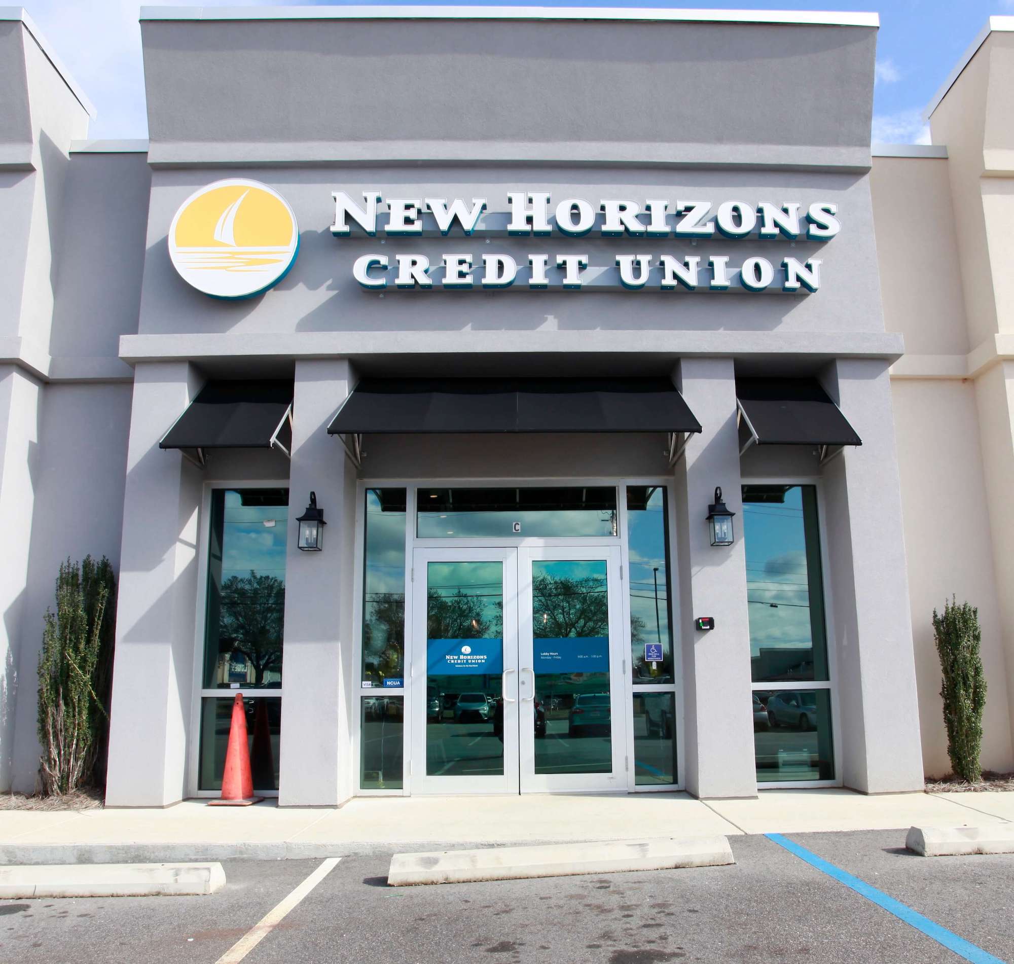 The exterior of a credit union storefront with light gray pillars, black window awnings, and three-dimensional signage reading “New Horizons Credit Union” in white, uppercase letters.