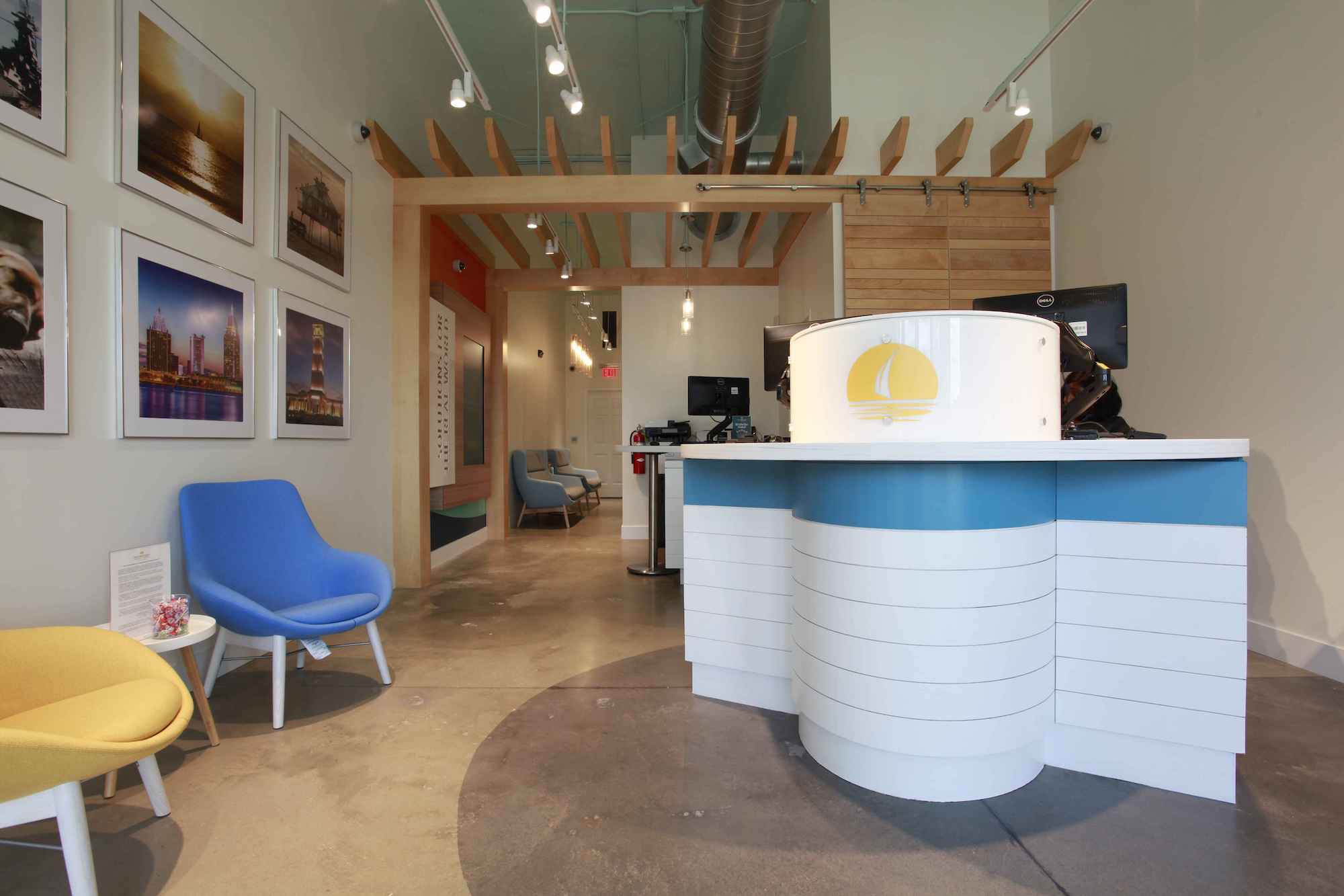 The lobby of New Horizons Credit Union at Rangeline Crossing. In the foreground is a white Teller Pod with a horizontal light blue stripe and a yellow logo of a sailboat on the front. The walls are white and feature framed photographs of local and oceanside scenes. Two chairs sit against one wall, one blue, the other yellow. The room extends back under an awning made of pale blond wood slats.