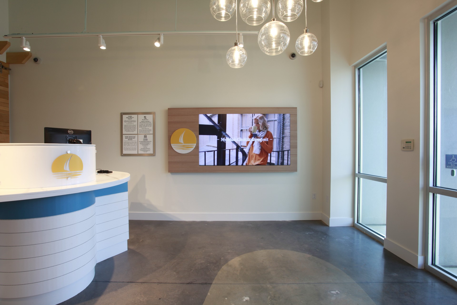 The Teller Pod and one wall of the New Horizons lobby. The Teller Pod is white with a light blue horizontal stripe and a yellow logo of a sailboat on the front. The wall is white and features a recessed video screen set into a panel of light blond wood. A chandelier made of clear glass globes hangs from the ceiling.