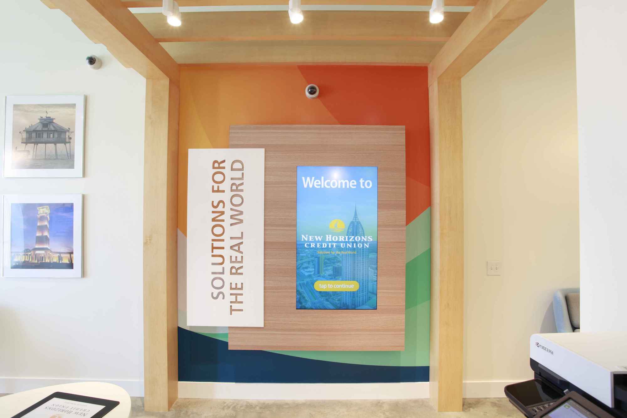 A wall featuring broad abstract stripes of orange, light green, and deep blue, a panel of blond wood, and a white sign reading “Solutions for the real world,” the New Horizons tagline. The wood panel contains a recessed video screen displaying the words “Welcome to New Horizons Credit Union” and a yellow button that says “Tap to continue.”