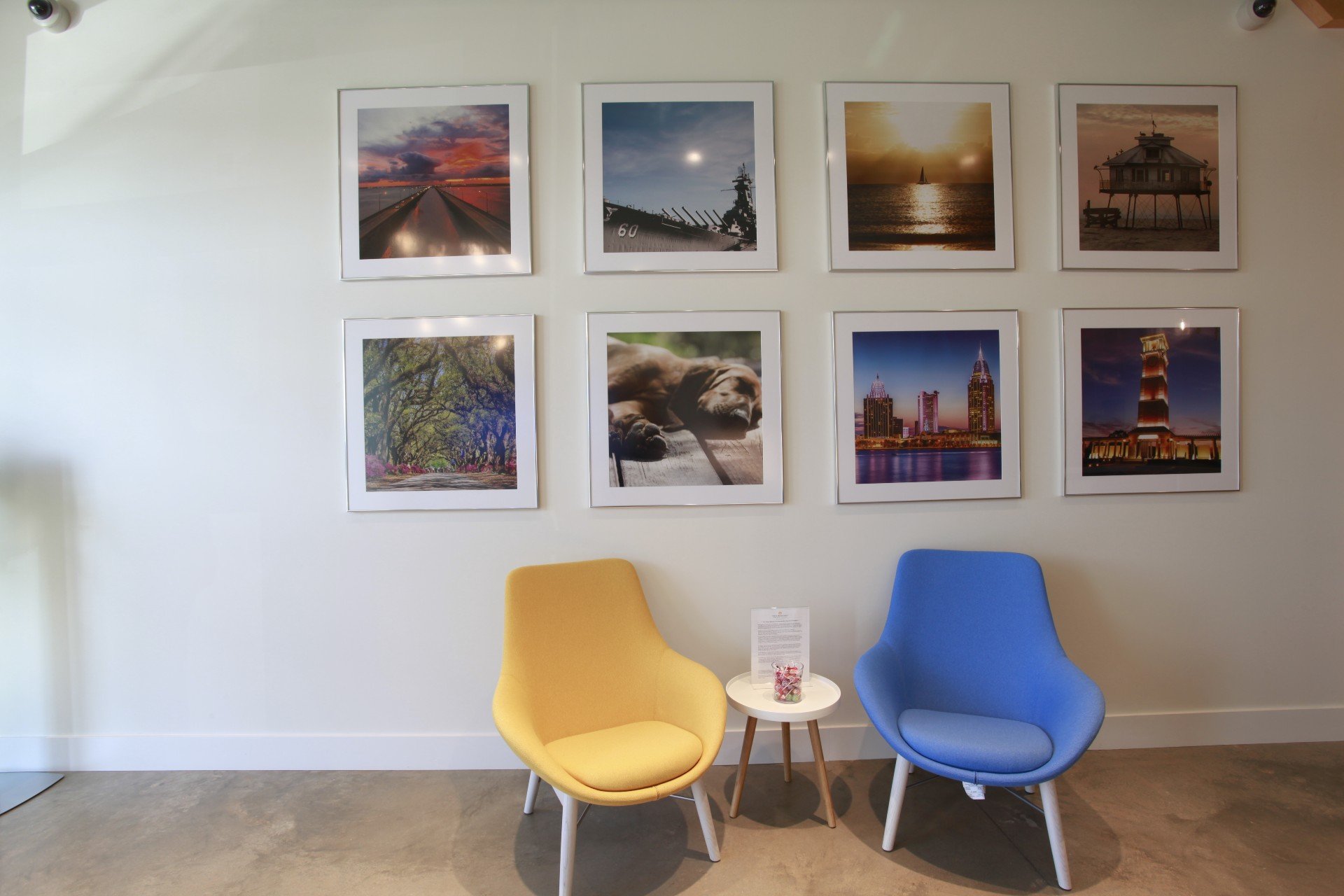 A white wall in the New Horizons lobby features two rows of framed photographs of local and oceanside scenes. Two chairs, one yellow and one blue, sit in front of the photographs with a small, round white table between them holding a jar of lollipops.