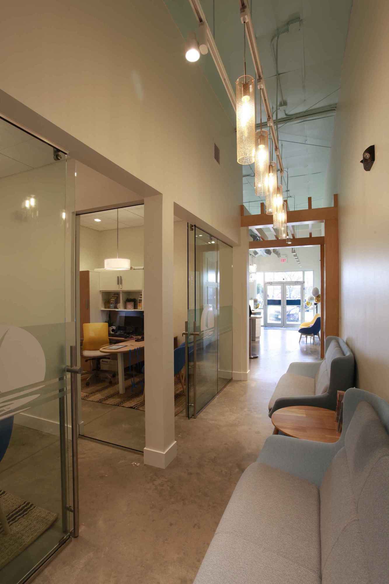 A glass-walled corner office containing a tan swivel chair and a desk, as viewed from the hallway outside. Two pale gray couches sit side by side along the hallway wall, and a row of slim glass pendant lights lines the ceiling. The front doors of the lobby can be seen beyond the hallway in the background.