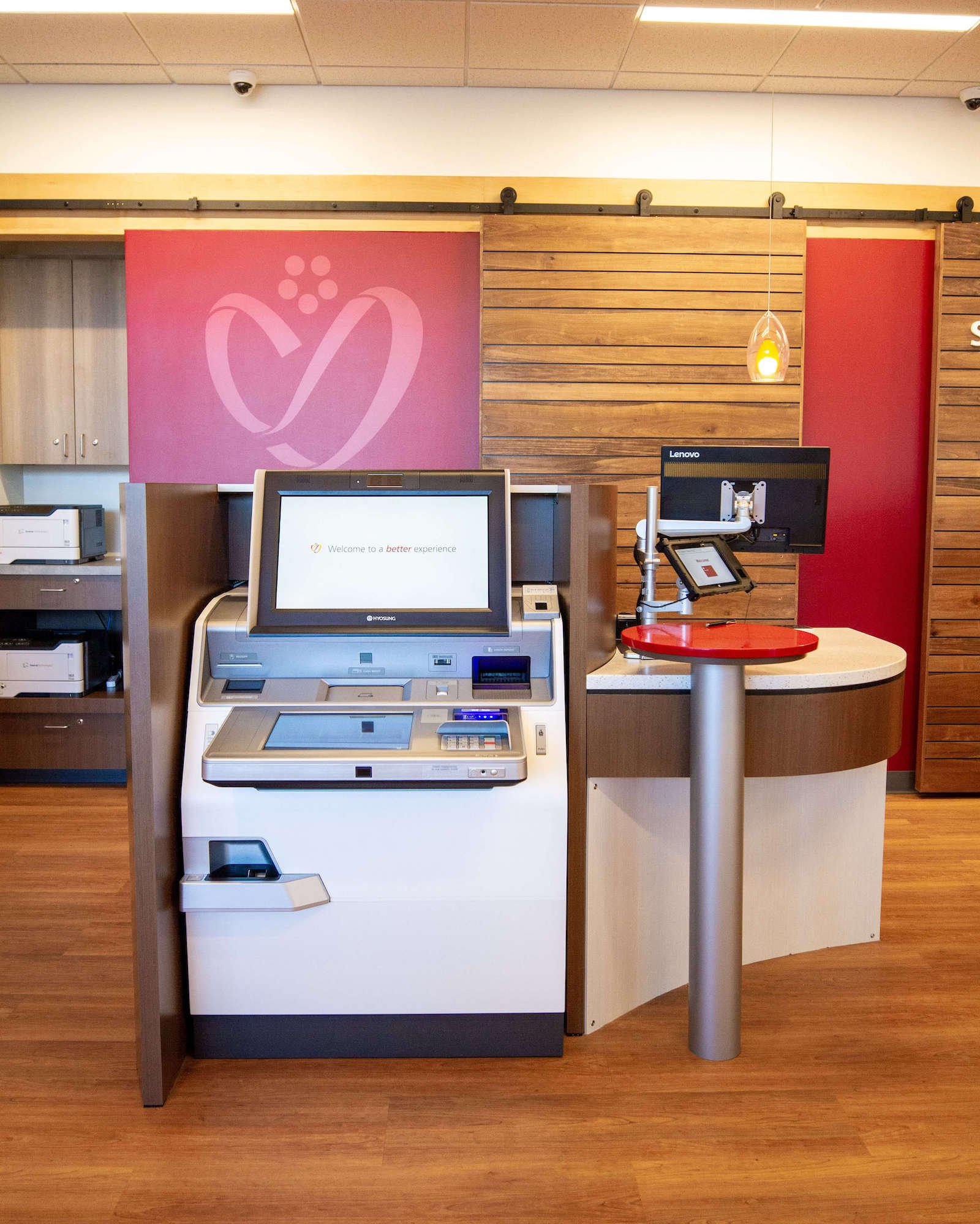 A credit union interior featuring a self-service kiosk with a touchscreen display. Behind the kiosk is a red wall with a heart-shaped logo against a wood panel wall.