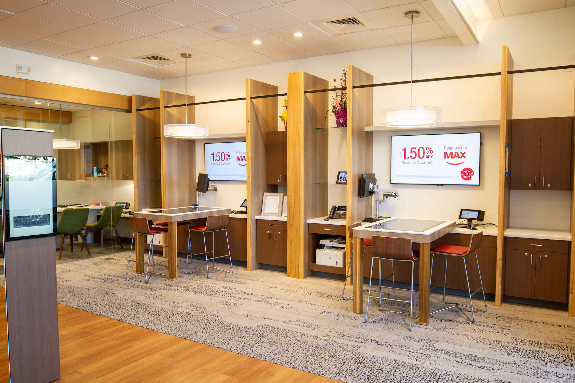 A modern credit union interior featuring a spacious layout with tall wooden partitions, a circular table with overhead pendant lighting, and a seating area with stools.