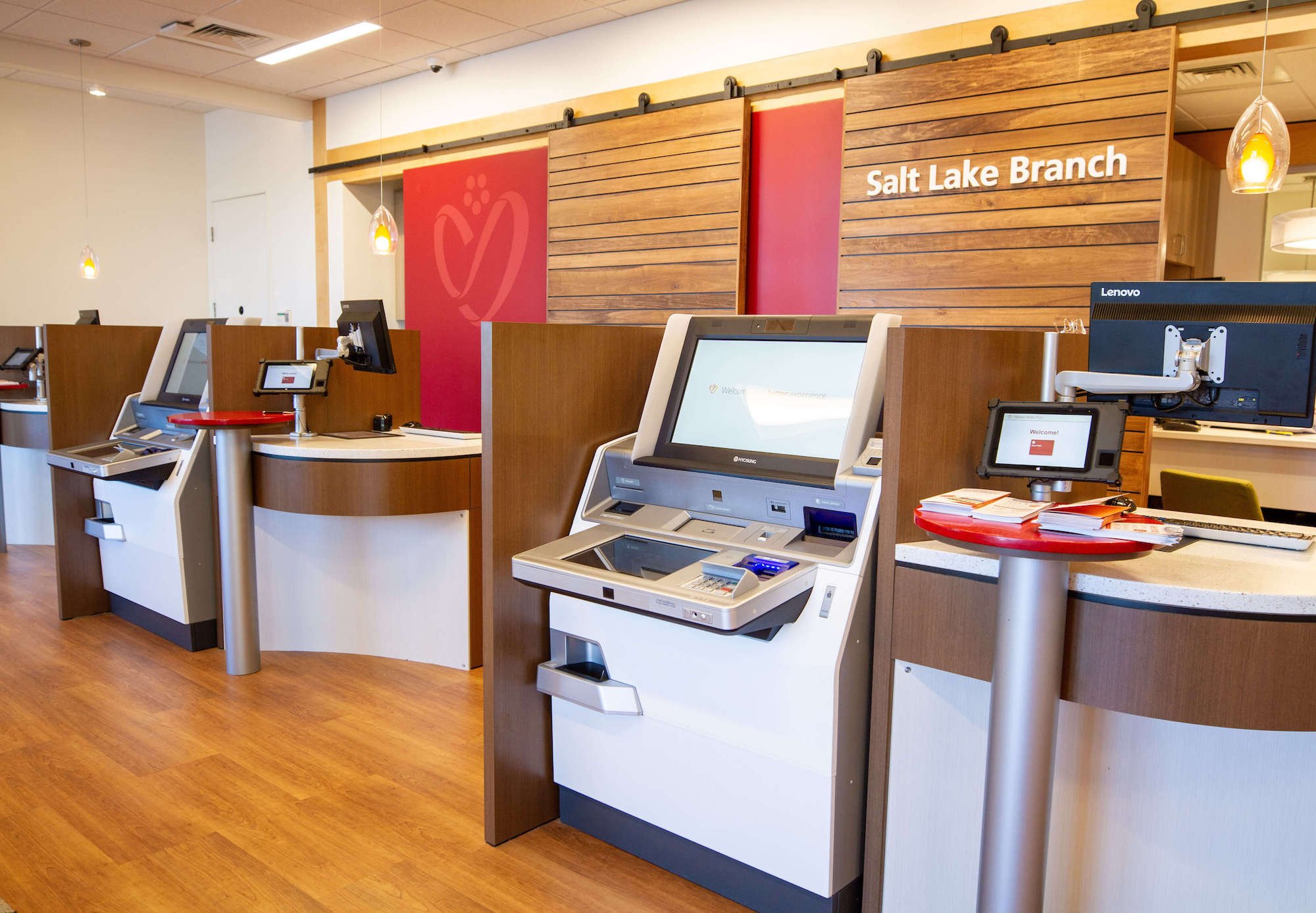 Interior of a credit union showcasing multiple self-service kiosks with touchscreen displays. There are computer workstations in the background.