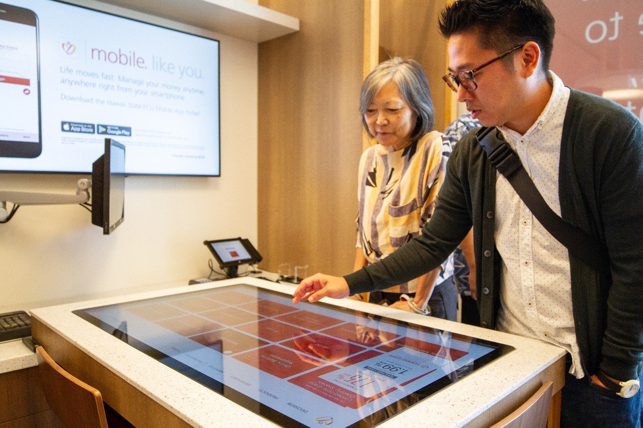 A woman and man stand side by side looking at an interactive touchscreen table. The man points at the screen. In the background, there's a wall-mounted screen.