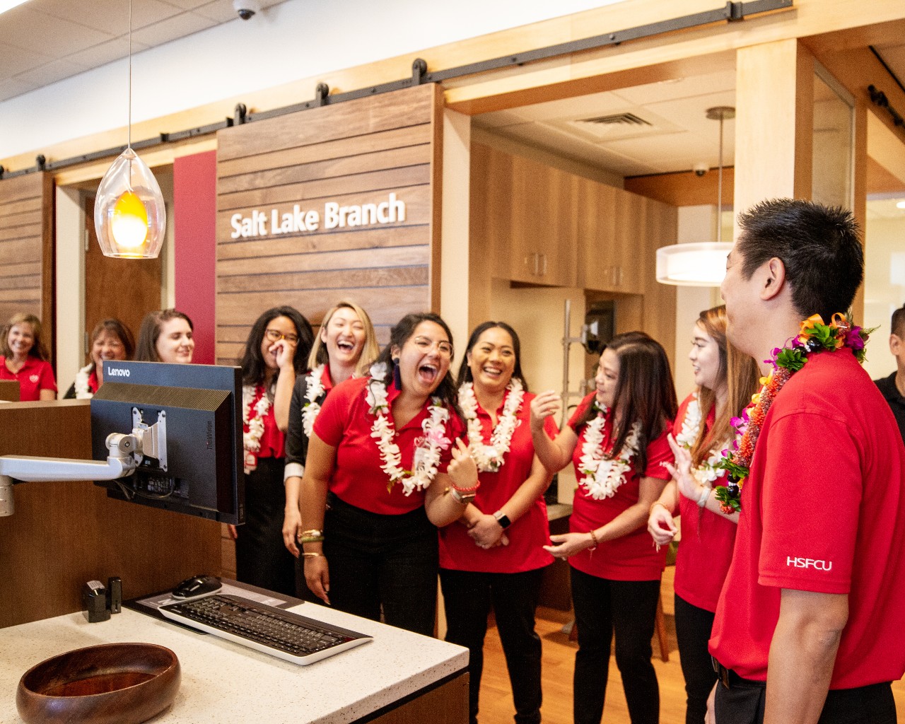 A group of credit union employees wearing red shirts and white floral leis laugh together, surrounded by a wood panel wall, a pendant light, and a computer workstation.