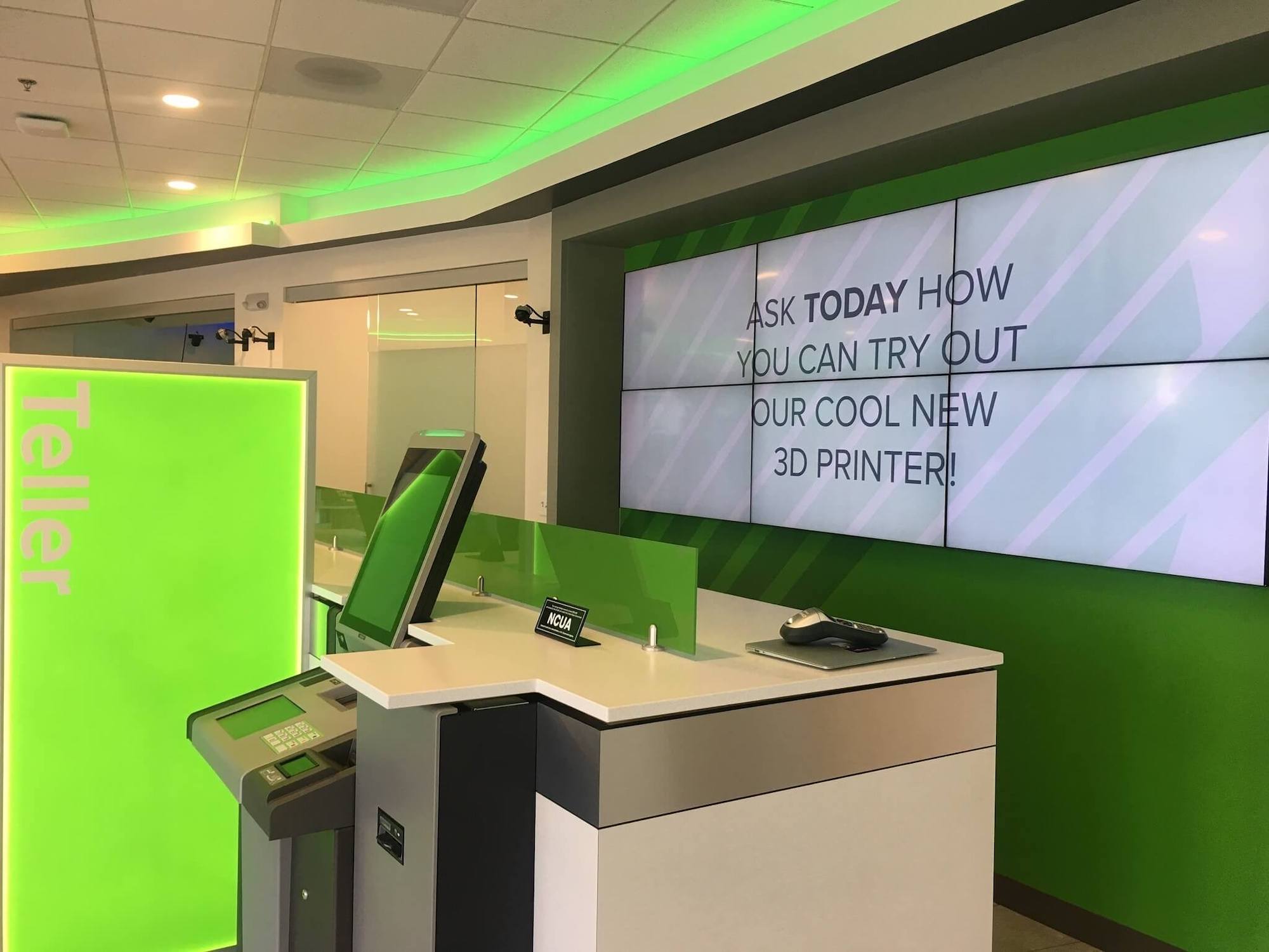 A modern teller counter with vibrant green lighting and a digital signage screen promoting the use of a new 3D printer available for customers.