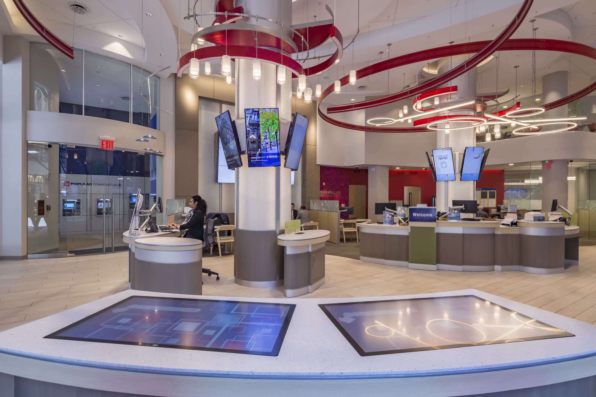 A financial institution interior featuring interactive digital counter displays, overhead digital signage, and a central seating area under stylish hanging lights.